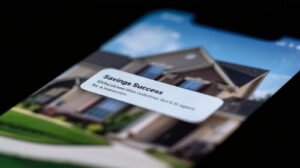 AI progress report 2026. Close-up of a smartphone screen showing a 'Savings Success' notification from an AI assistant with a blurred US suburban home in the background.